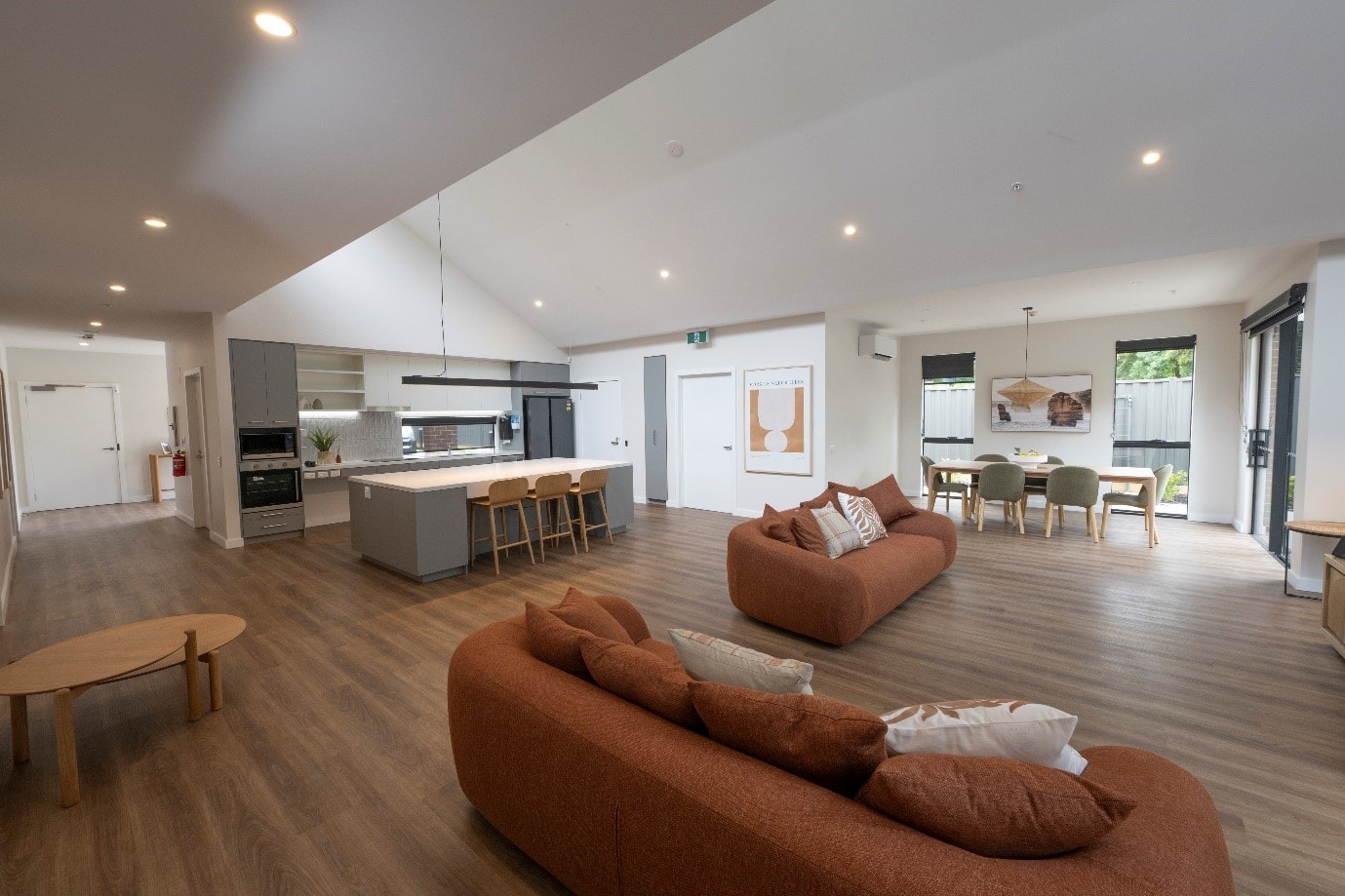 Open plan living area of Eltham house 2 with brown-orange couches in foreground, dining table with six chairs in the background and kitchen to the left.